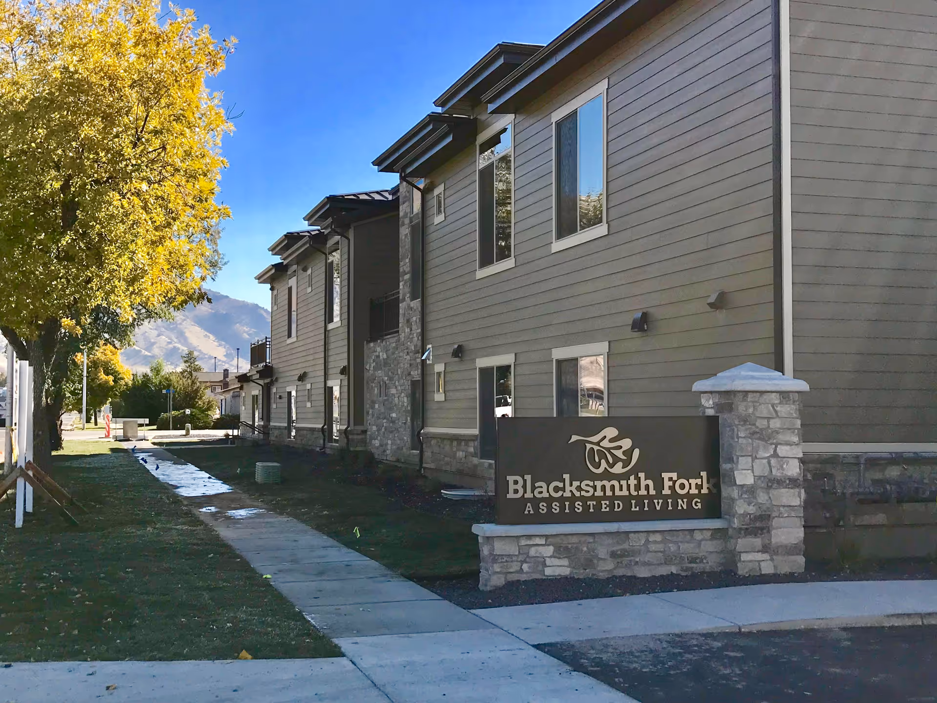 Exterior view of Blacksmith Fork Assisted Living facility showing a modern multi-story building with beige siding and stone accents. A sidewalk runs alongside the building with a tree on the left side and mountains visible in the background under a clear blue sky.