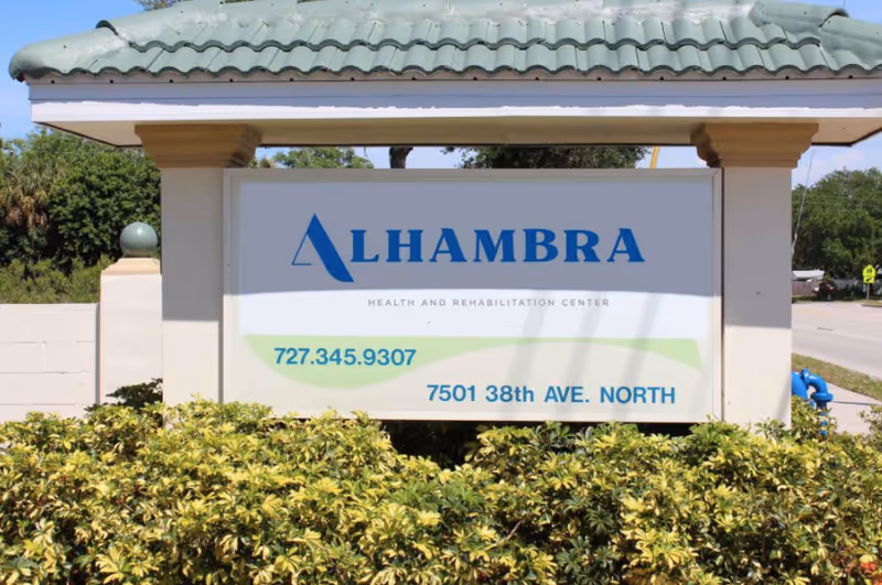 Outdoor sign for Alhambra Health and Rehabilitation Center with the phone number 727.345.9307 and address 7501 38th Ave. North, surrounded by green bushes and a tiled roof structure above the sign.