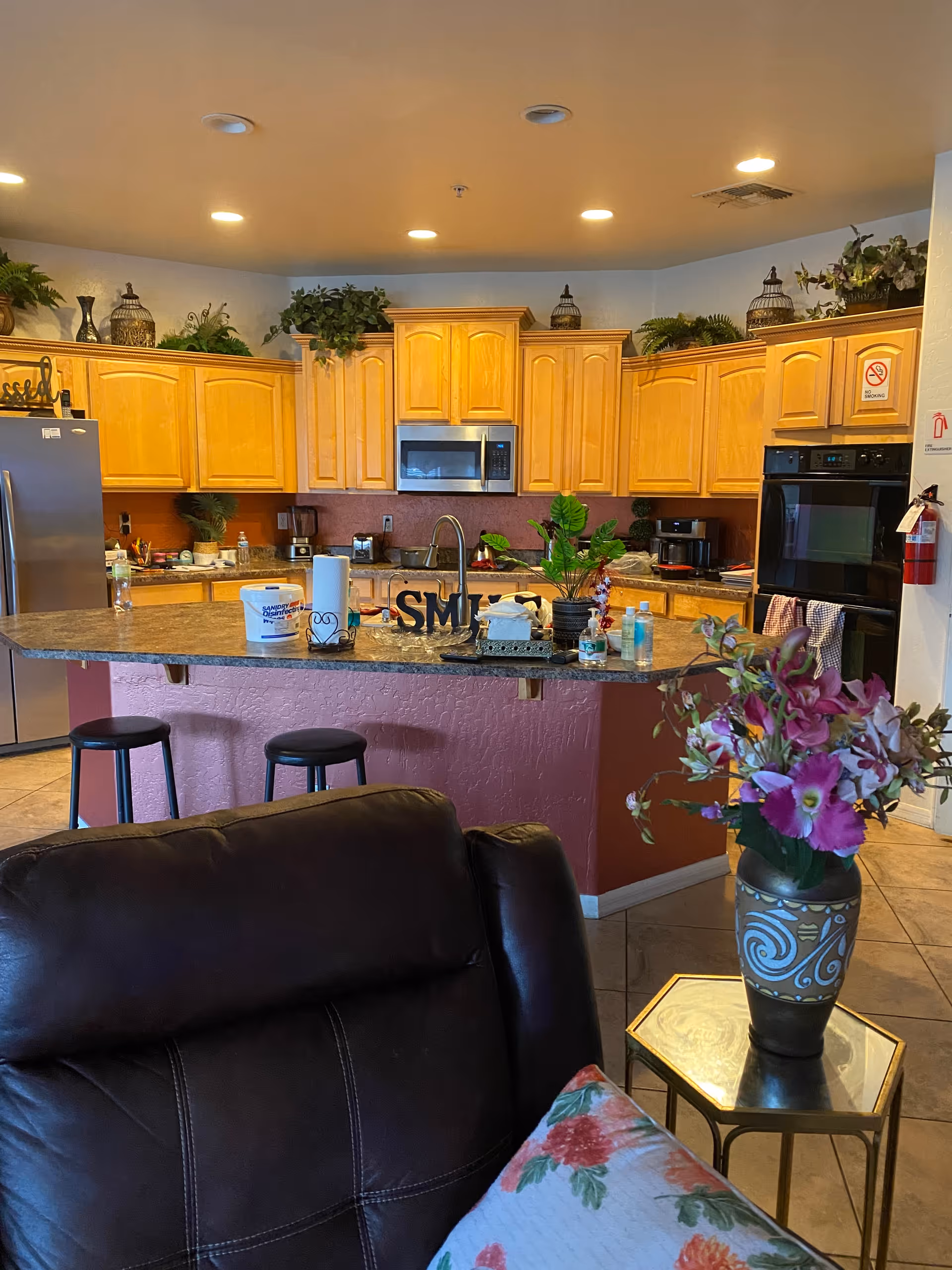 Interior view of a kitchen with wooden cabinets, a granite countertop island with two black stools, and various kitchen appliances including a microwave and oven. There are decorative plants on top of the cabinets and a vase with purple flowers on a small side table next to a dark brown leather chair with a floral pillow.