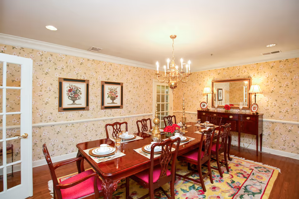 A traditional dining room with a polished wooden dining table set for eight people with white plates, cups, and glasses. The chairs have red cushions. The room features floral wallpaper, a colorful floral rug, a chandelier hanging above the table, two framed floral paintings on one wall, and a wooden sideboard with a large mirror and two lamps on the opposite wall.