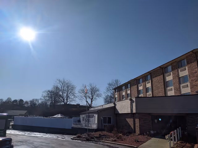 Exterior view of a multi-story brick building under a clear blue sky with the sun shining brightly. There are leafless trees and a flagpole with an American flag in front of the building. The ground is partially covered with snow and there is a paved driveway or parking area in front.