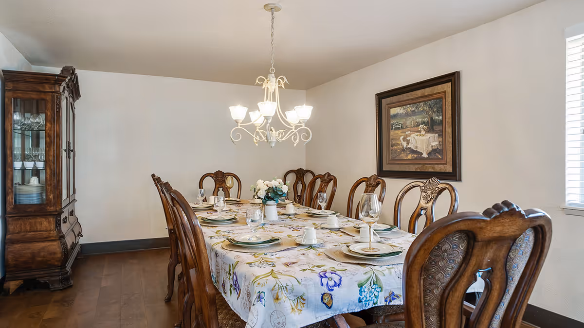 A dining room with a long wooden table covered with a floral tablecloth set with plates, cups, and glasses. The room features wooden chairs with ornate backs, a chandelier hanging above the table, a wooden china cabinet with glassware on the left, and a framed painting on the wall to the right.
