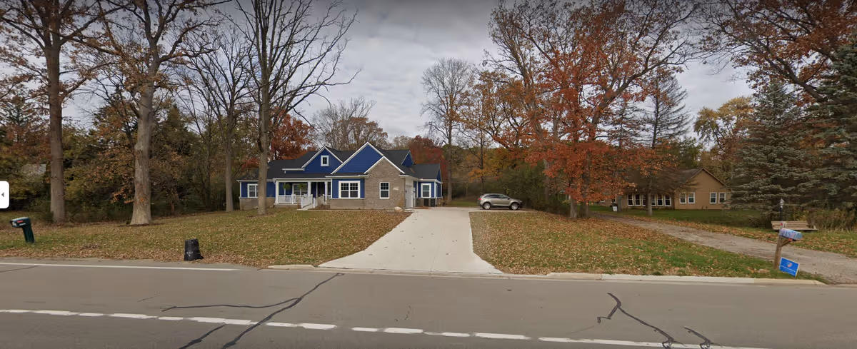 A single-story residential building with a blue roof and beige stone facade, surrounded by trees with autumn foliage. There is a concrete driveway leading up to the house, a car parked on the right side, and a mailbox near the street. The scene is set on a cloudy day.