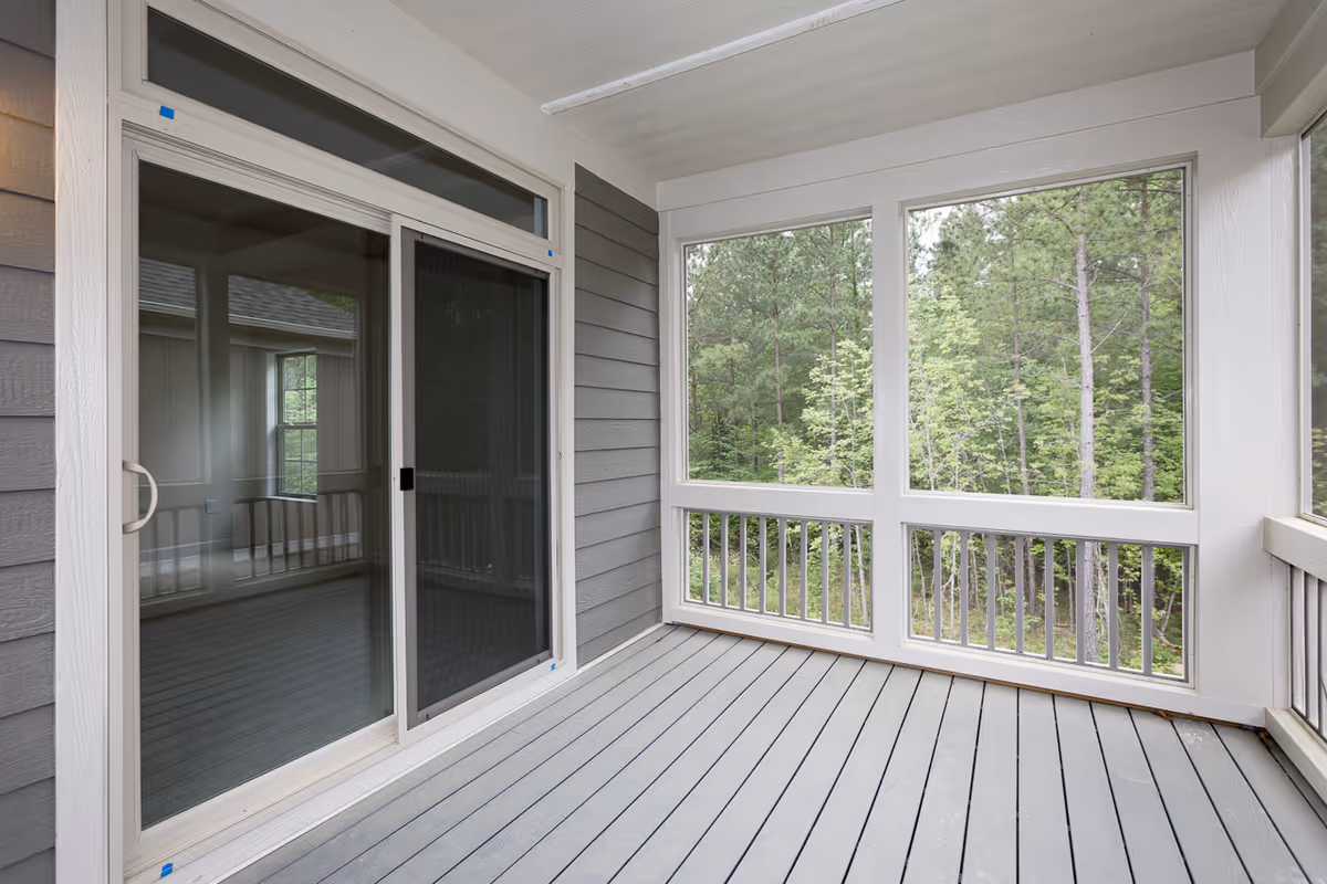 A screened-in porch with gray wooden flooring and white framing. The porch has large screened windows overlooking a green wooded area. There is a sliding glass door leading inside the building.