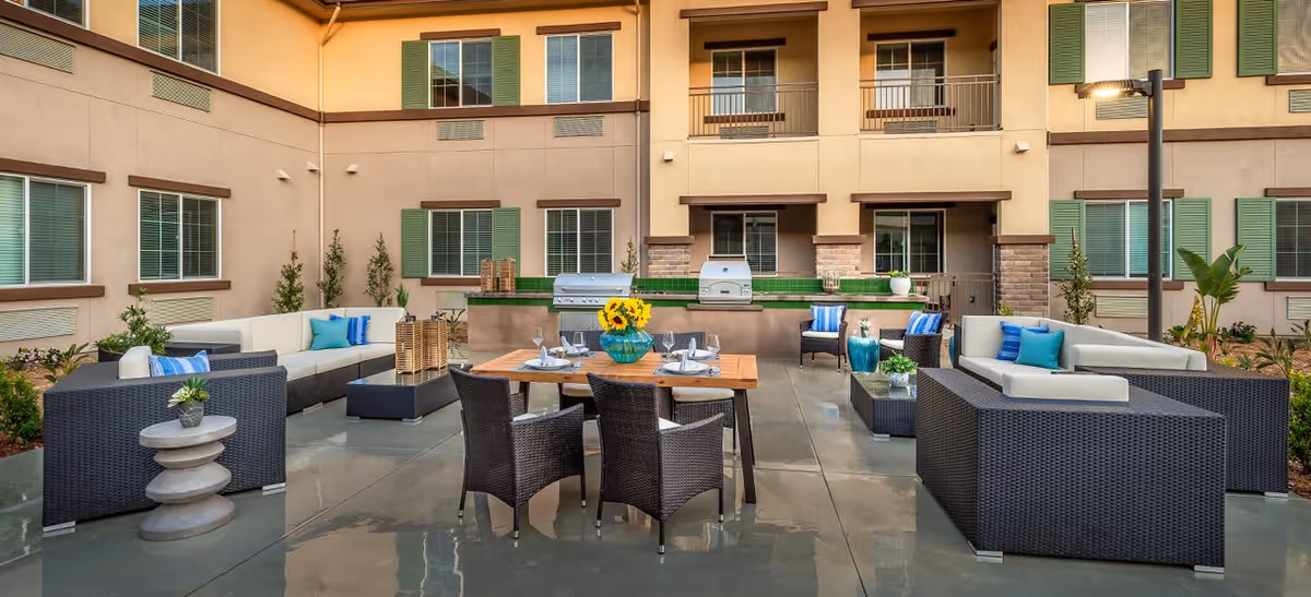 Outdoor patio area at Ocean Hills Senior Living featuring modern wicker furniture with white cushions, a wooden dining table set with plates and glasses, decorative plants, and a built-in barbecue grill against the building wall with windows and green shutters.