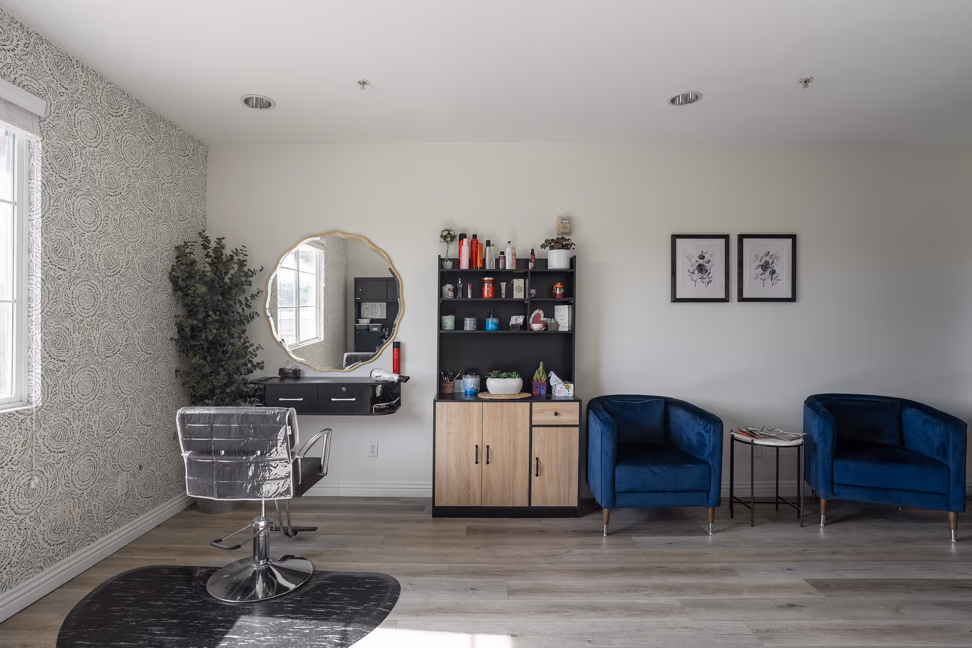 A well-lit room with a styling chair in front of a round mirror mounted on a wall with patterned wallpaper. Next to the mirror is a black and wood cabinet filled with various hair and beauty products. To the right, there are two blue armchairs with a small round table between them, holding magazines. The floor is light wood, and the walls are white with two framed floral artworks above the chairs.