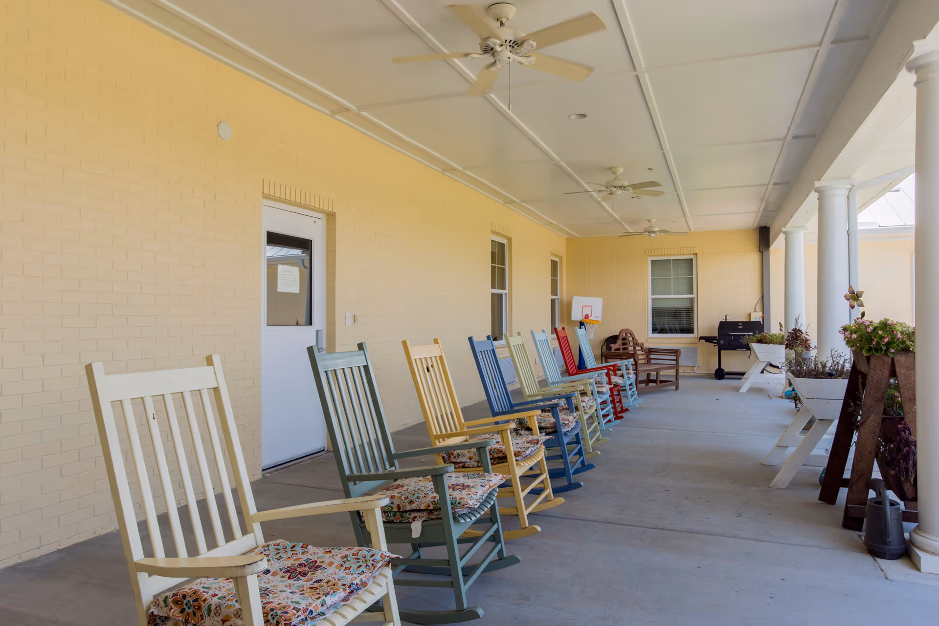 Covered outdoor patio area with a row of colorful rocking chairs with floral cushions lined up against a yellow brick wall. Ceiling fans are mounted on the white ceiling, and there are white columns on the right side. A small basketball hoop, a wooden bench, a grill, and several planters with flowers are visible in the background.