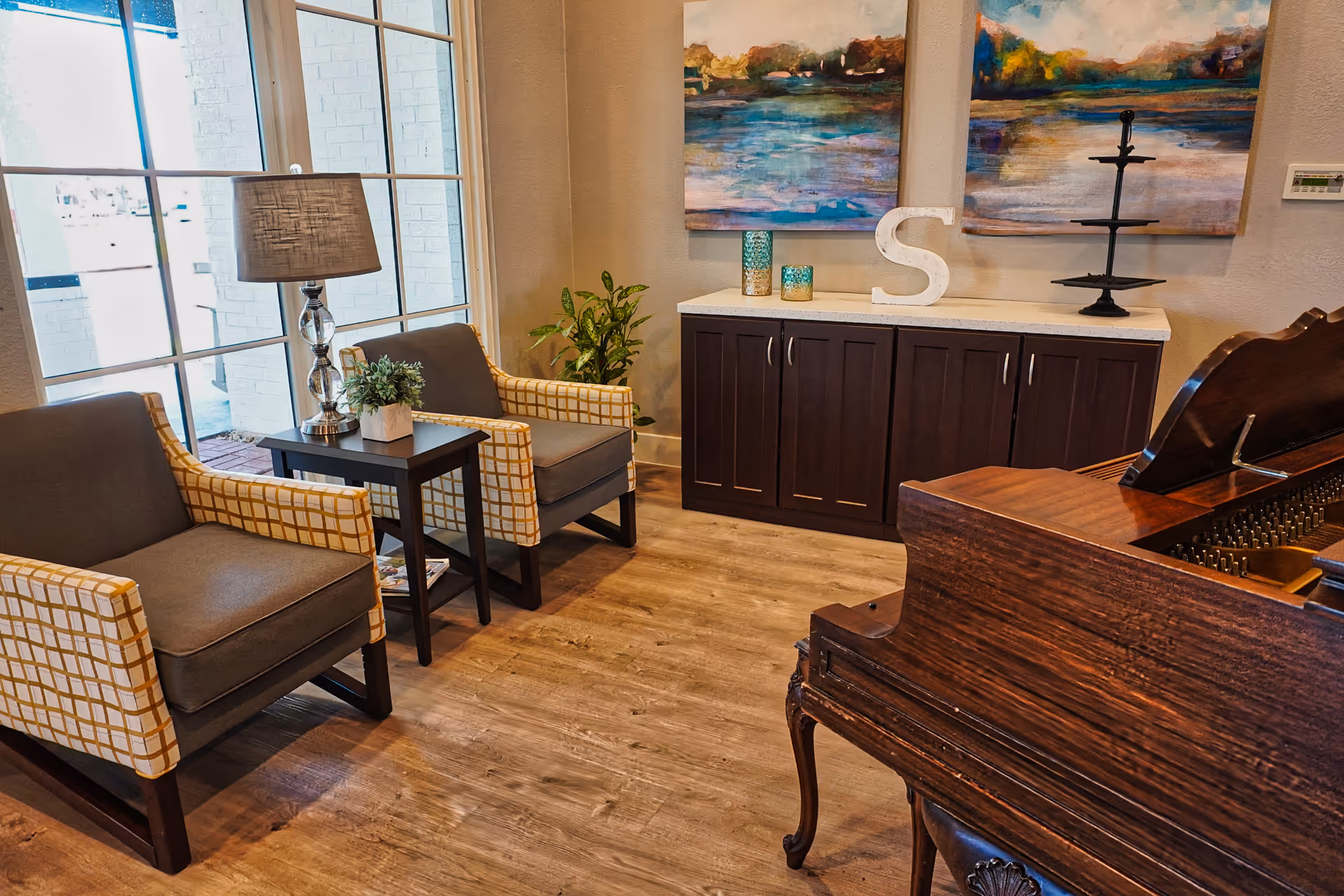 A cozy interior room with two armchairs featuring yellow and white checkered sides and gray cushions, a small black side table with a lamp and a potted plant, a dark wooden cabinet with decorative items including a large letter 'S' and two abstract paintings on the wall, and a wooden piano partially visible in the foreground.