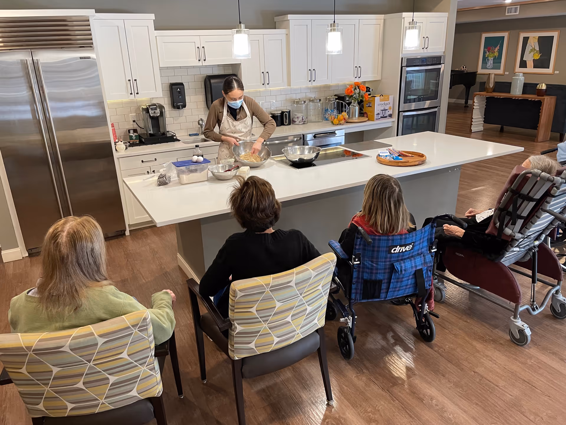 A woman wearing a mask and apron is preparing food in a modern kitchen with white cabinets and stainless steel appliances. Four elderly individuals, two seated in chairs and two in wheelchairs, watch her from the opposite side of a large kitchen island.