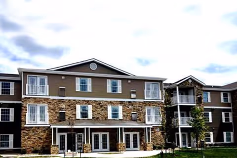 Three-story retirement community building with stone and siding facade, balconies, and a covered entry under a cloudy sky.
