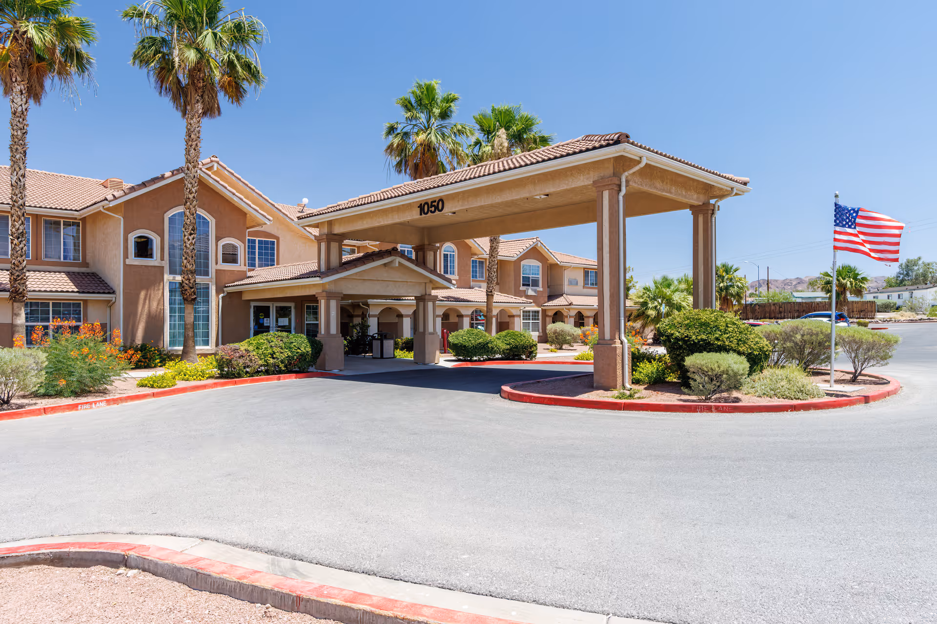 Front entrance of a stucco assisted living building with a covered porte-cochère, palm trees, landscaped beds, and an American flag.