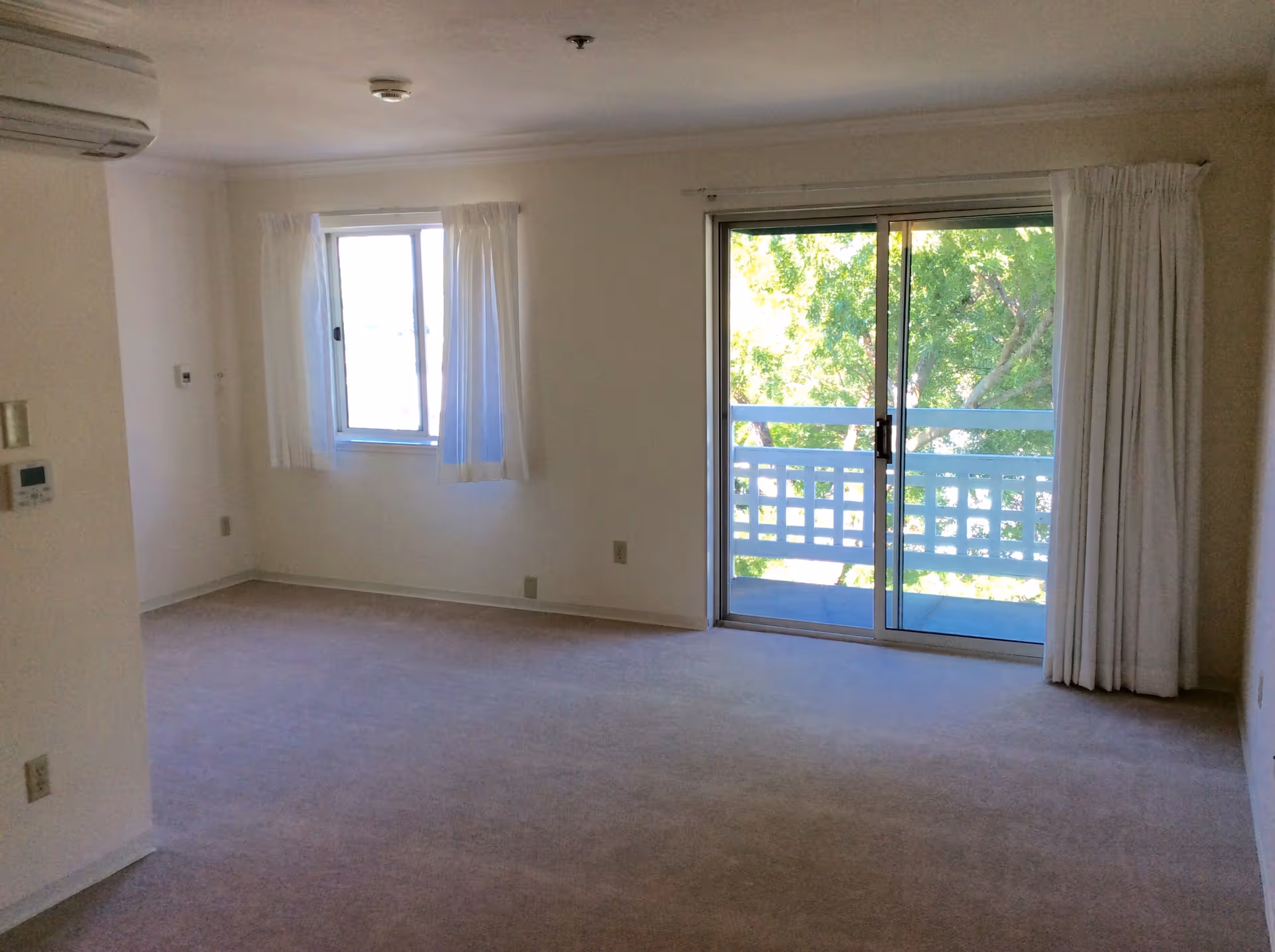 Empty bright living room with a sliding glass door to a balcony, a curtained window, and beige carpeting.