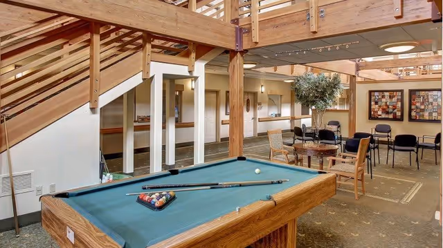 Interior view of a senior living facility common area featuring a pool table with cues and balls arranged, wooden staircase with railing, several chairs and tables, a potted tree, and framed artwork on the walls under a ceiling with recessed lighting.