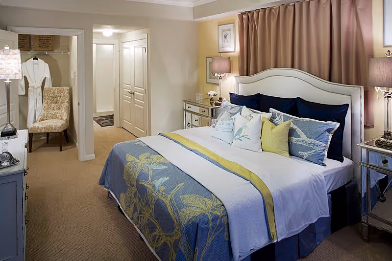 A well-decorated bedroom featuring a large bed with multiple pillows and a blue and yellow floral bedspread. The room includes two mirrored nightstands with lamps, a chair near an open closet with a hanging white robe, and a hallway with closed double doors in the background. The walls are painted in soft neutral tones with a curtain behind the bed.