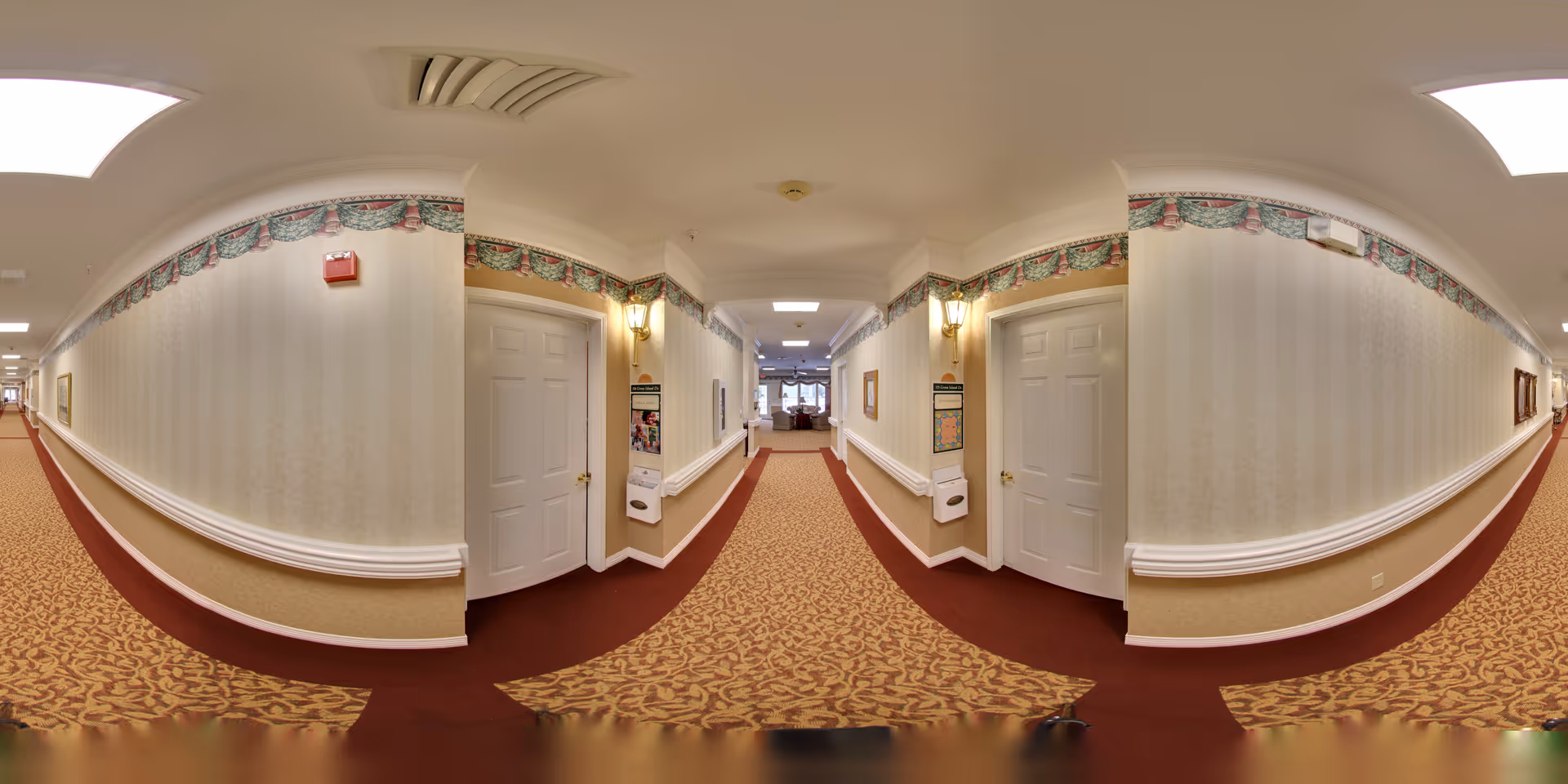 Long carpeted interior hallway in a senior living facility with patterned wallpaper, doors on both sides and a seating area visible at the far end.