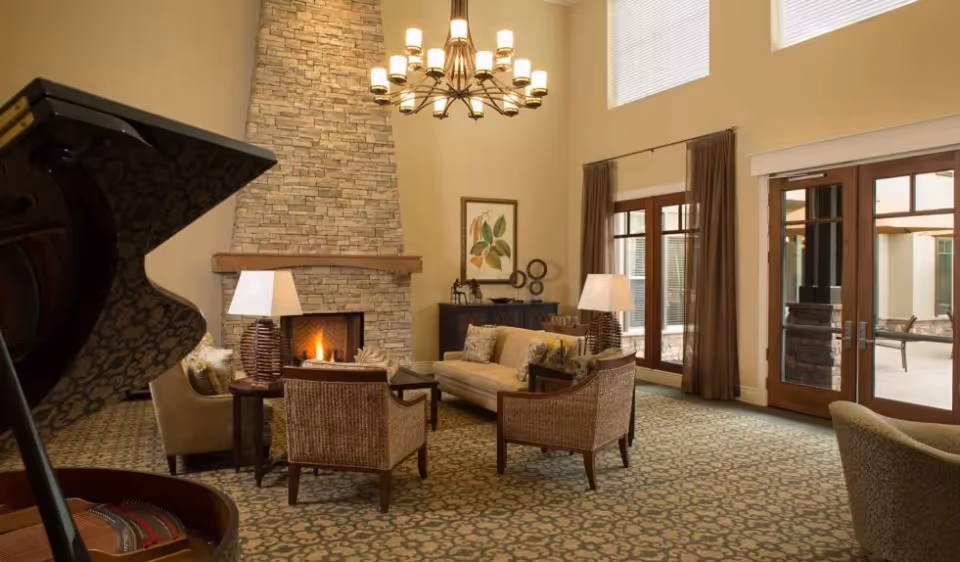 Spacious senior living common room with a stone fireplace, seating area, grand piano, chandelier, and glass doors to a patio.