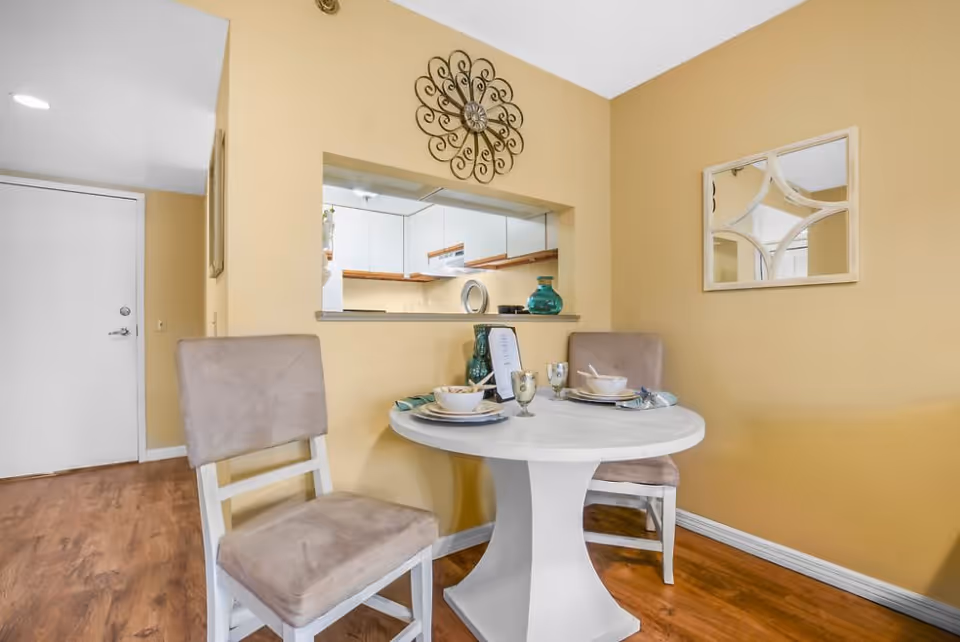 A small dining area with a round white table set for two, featuring beige cushioned chairs. The wall behind the table is painted yellow and decorated with a decorative metal wall art and a mirror. A pass-through window reveals a white kitchen with wooden accents. The floor is wooden, and a white door is visible in the background.