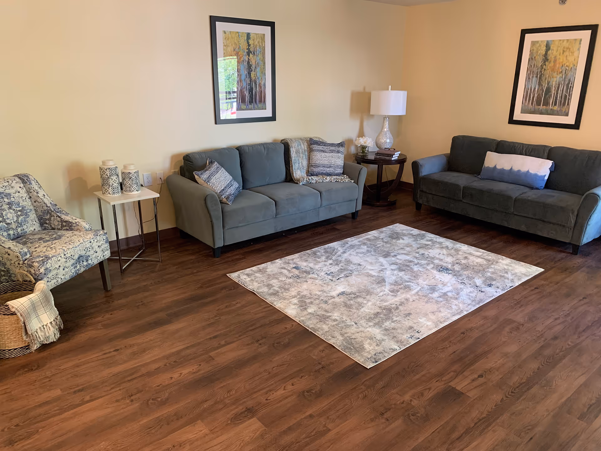 A cozy living room with two gray sofas, a patterned armchair, a small white side table with decorative vases, a round wooden side table with a lamp and flowers, two framed paintings of trees on the walls, and a light-colored area rug on a wooden floor.