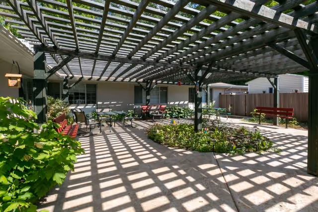 Outdoor patio area with a wooden pergola casting shadows on the concrete floor, surrounded by green plants and red benches. The patio is adjacent to a building with windows and a fenced backyard.
