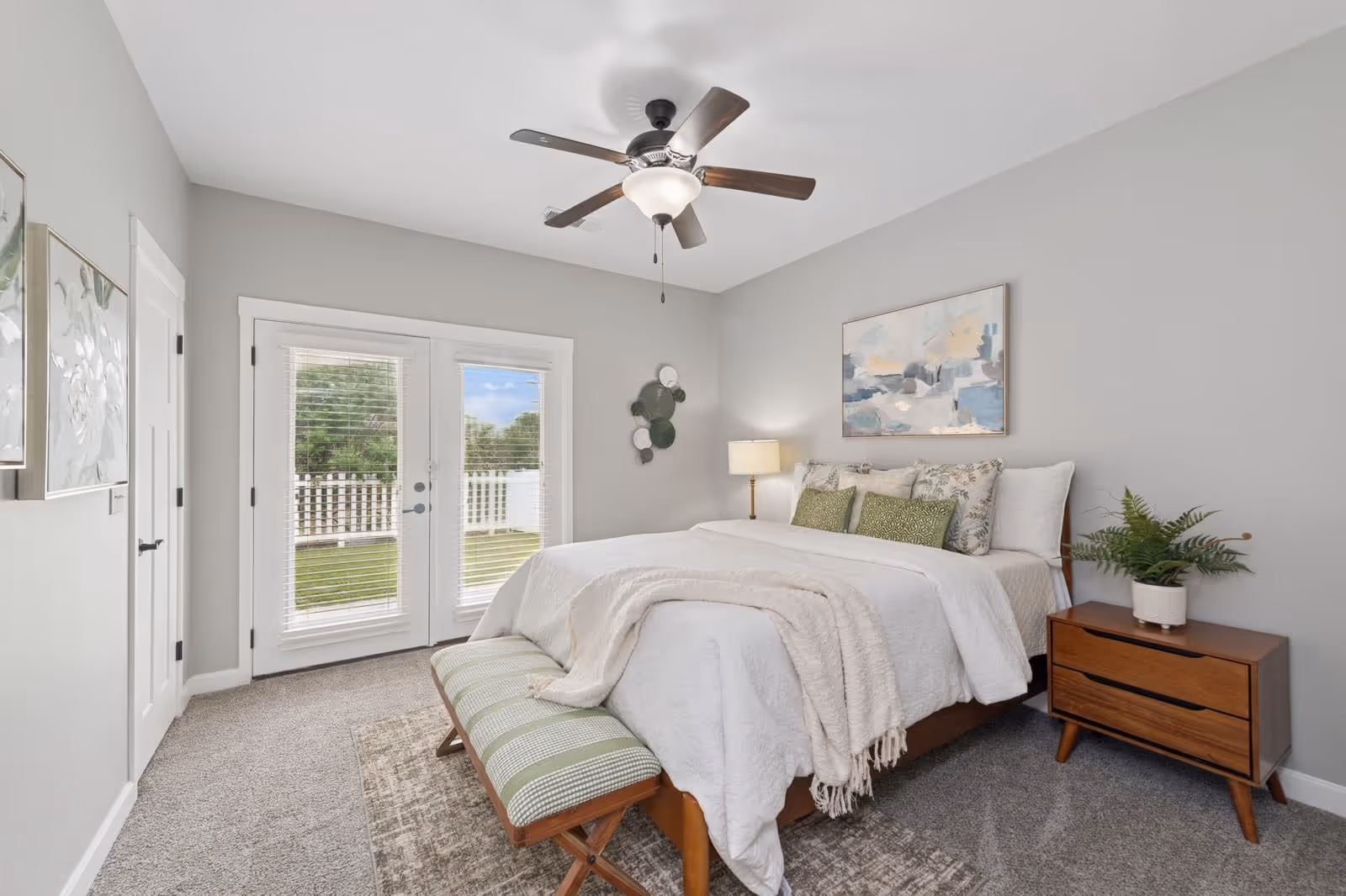 A bright bedroom with a large bed covered in white bedding and green accent pillows. There is a wooden bench with a green cushion at the foot of the bed, a wooden nightstand with a potted plant and a lamp, and a ceiling fan with lights. French doors with blinds lead outside to a fenced yard. The walls are painted light gray and decorated with framed artwork.