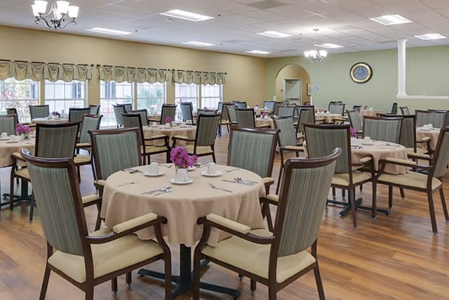 Well-lit dining room with round tables covered in beige tablecloths, floral centerpieces, and upholstered chairs.