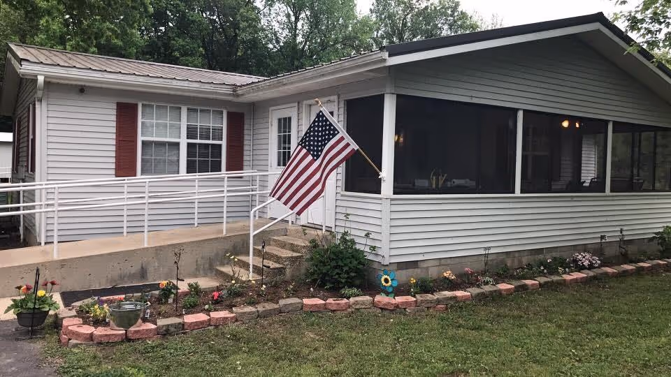 Exterior view of a single-story residential building with white siding and red shutters. There is a concrete ramp with white railings leading to a door, and an American flag is mounted on a pole near the steps. A flower bed with various plants and flowers is bordered by red bricks in front of the building. Trees are visible in the background.