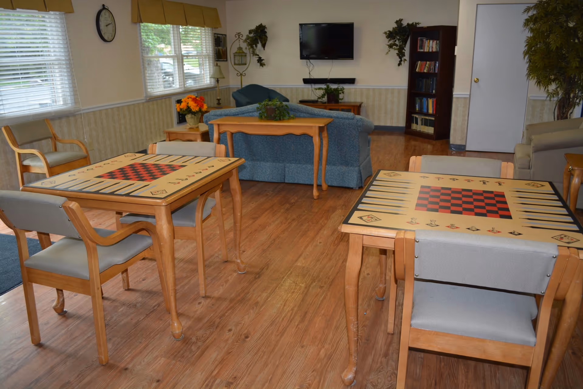 A cozy common area in a senior living facility with two wooden tables featuring checkerboard and backgammon game designs, surrounded by chairs. In the background, there are blue couches, a wall-mounted TV, a bookshelf, and decorative plants. The room has wooden flooring and windows with blinds allowing natural light.