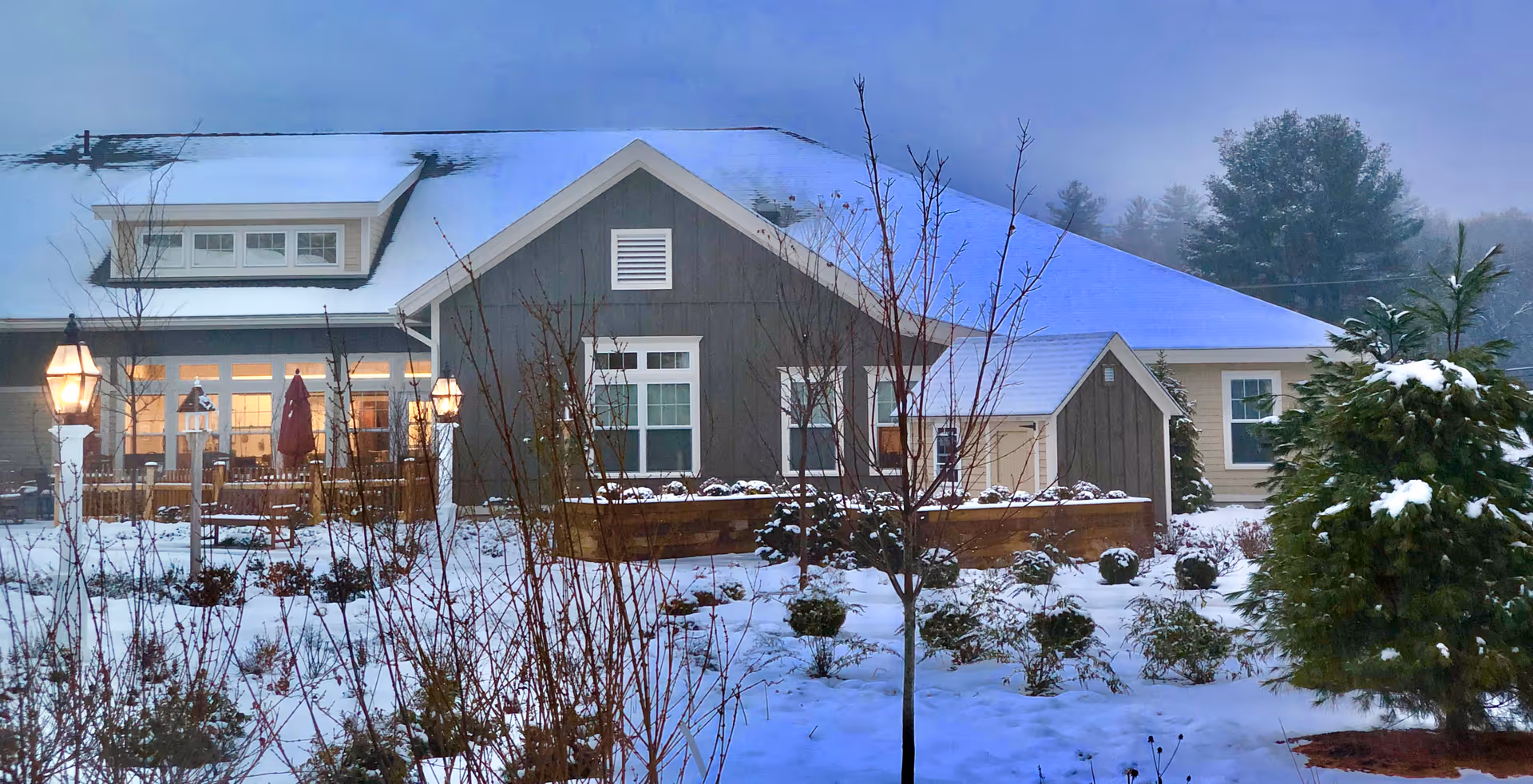 Exterior view of a senior living facility building covered with snow. The building has a gray and beige facade with multiple windows and a snow-covered roof. There are leafless trees and shrubs in the snowy garden area in front of the building, and two lit lamp posts near a patio with outdoor furniture.