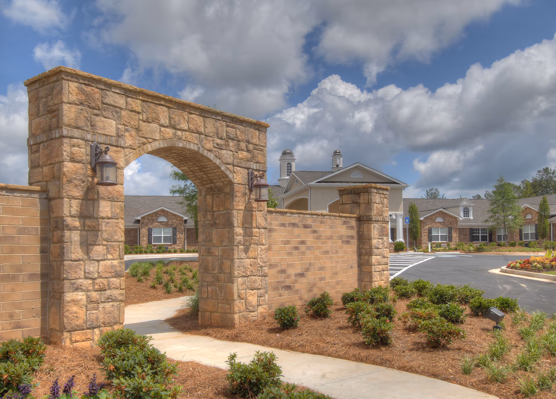 Stone archway entrance with lantern-style lights on each side, leading to a landscaped pathway with bushes and mulch. In the background, there is a beige building with white trim, cupolas on the roof, and a parking area with marked spaces. The sky is partly cloudy.