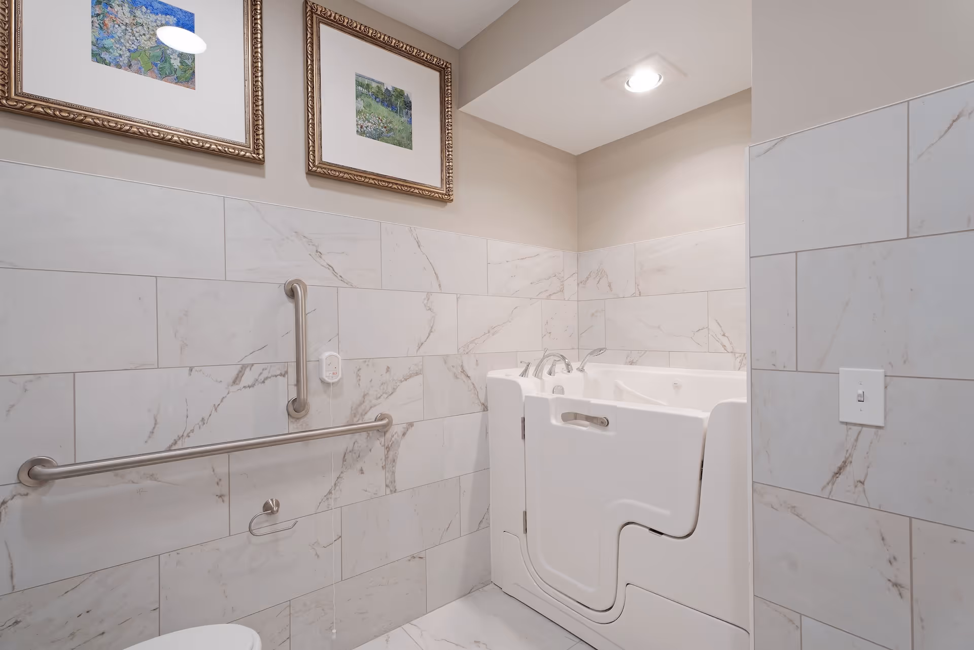 Accessible bathroom with a walk-in bathtub, stainless grab bars, marble tile walls, and framed artwork.
