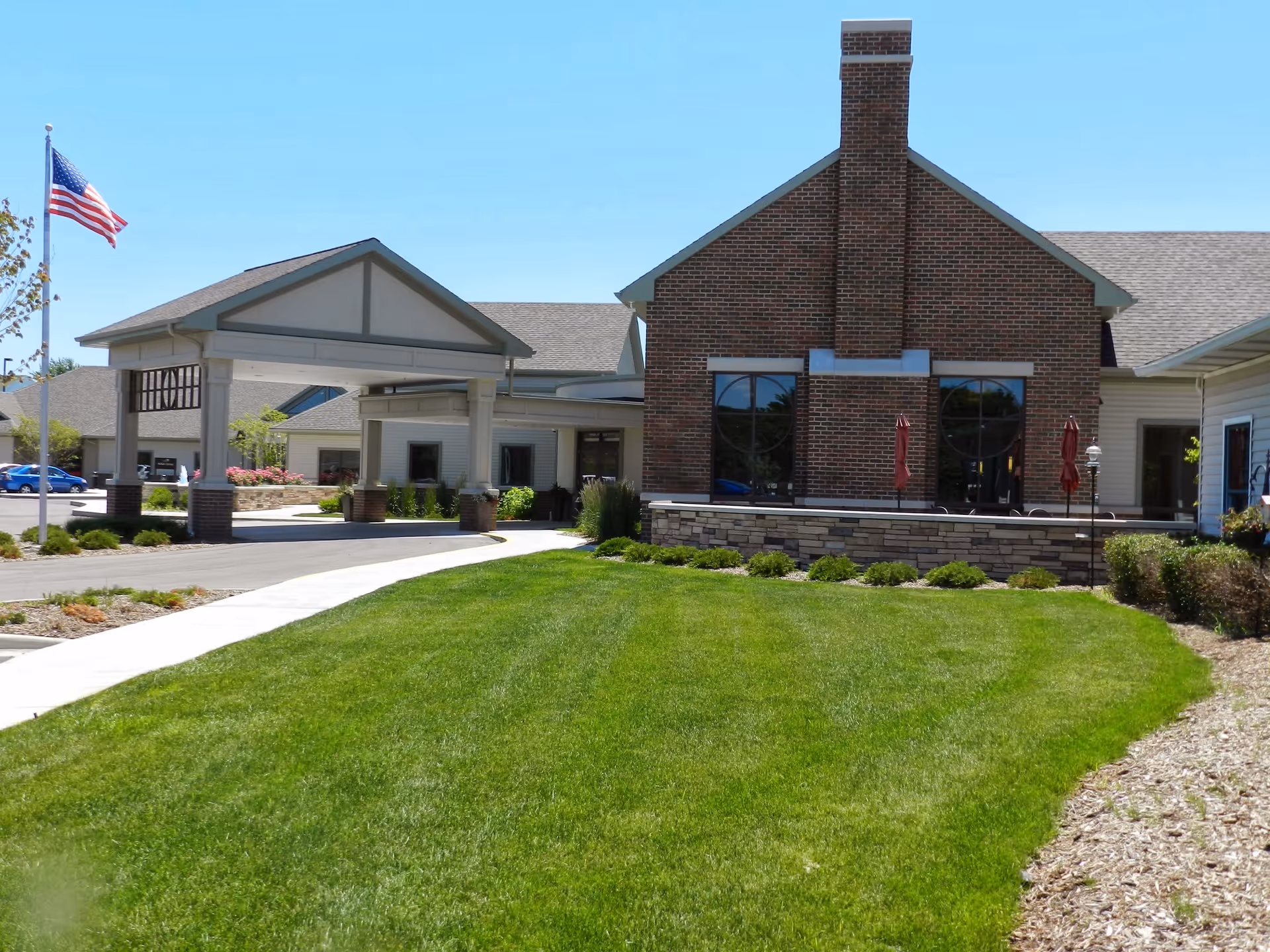 Exterior view of Resthaven Care Center showing a well-maintained green lawn, a brick building with large windows, a covered entrance with columns, and an American flag on a flagpole under a clear blue sky.