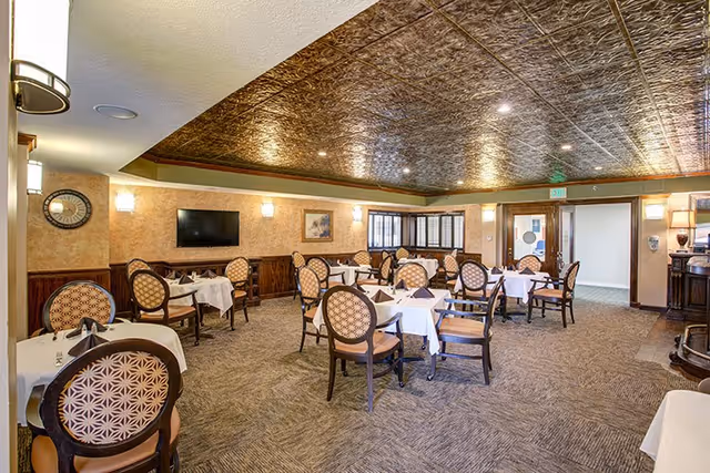 Formal dining room with multiple tables set with white tablecloths and patterned chairs under a decorative tin ceiling.