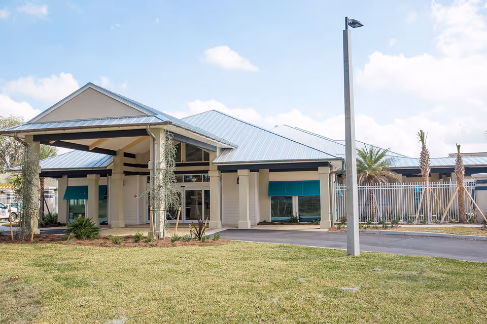 Exterior view of a single-story assisted living facility building with a metal roof, large covered entrance, green window awnings, palm trees, and a grassy lawn under a partly cloudy sky.