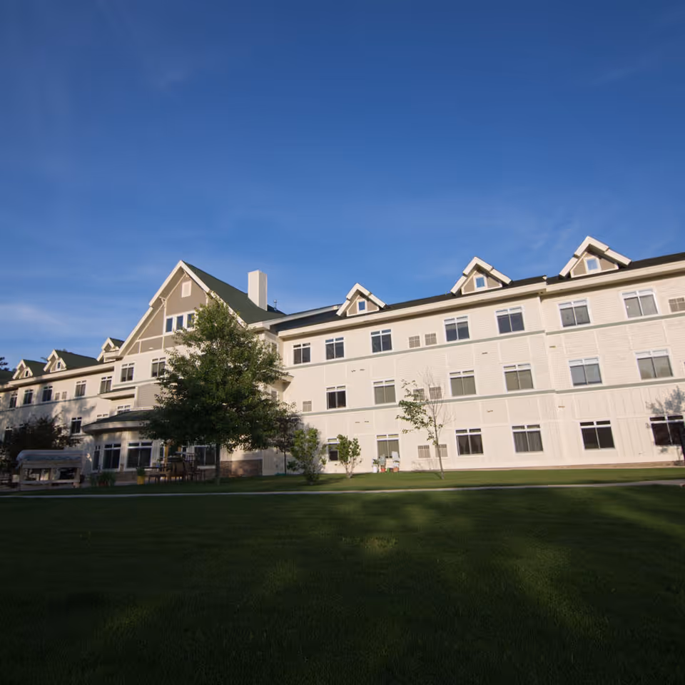 Exterior view of a large, multi-story assisted living facility building with multiple windows, a green lawn in the foreground, and a clear blue sky above.