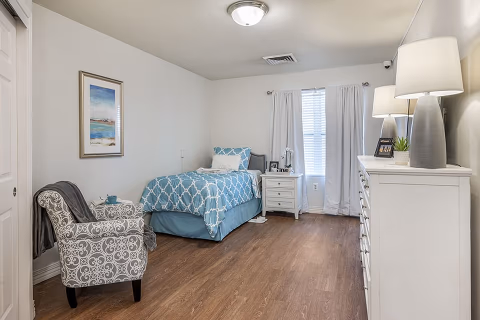 A neatly arranged bedroom with a single bed covered in a blue and white patterned bedspread, a white nightstand with a lamp and decorative items, a white dresser with two lamps and a small plant, a patterned armchair with a gray throw draped over it, a framed artwork on the wall, and a window with white curtains allowing natural light to enter.