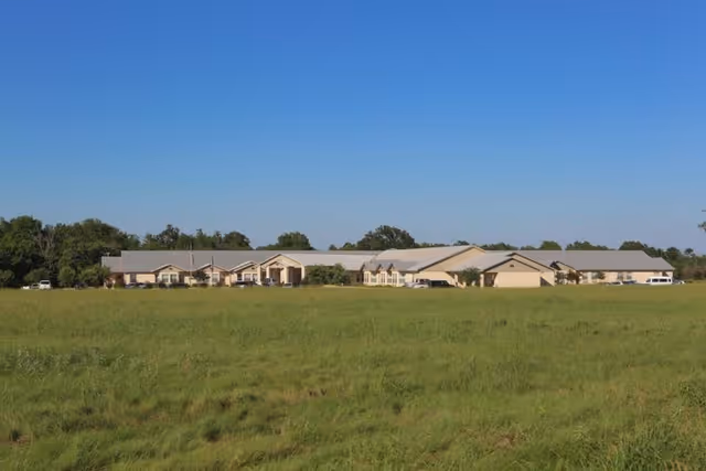 Wide view of a single-story rehabilitation and healthcare center building with a light-colored roof, situated behind a large grassy field under a clear blue sky.