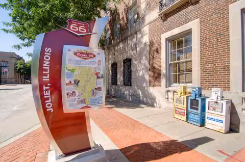 A Route 66 informational sign labeled 'Joliet, Illinois' stands on a brick sidewalk beside a brick building and a row of newspaper boxes.
