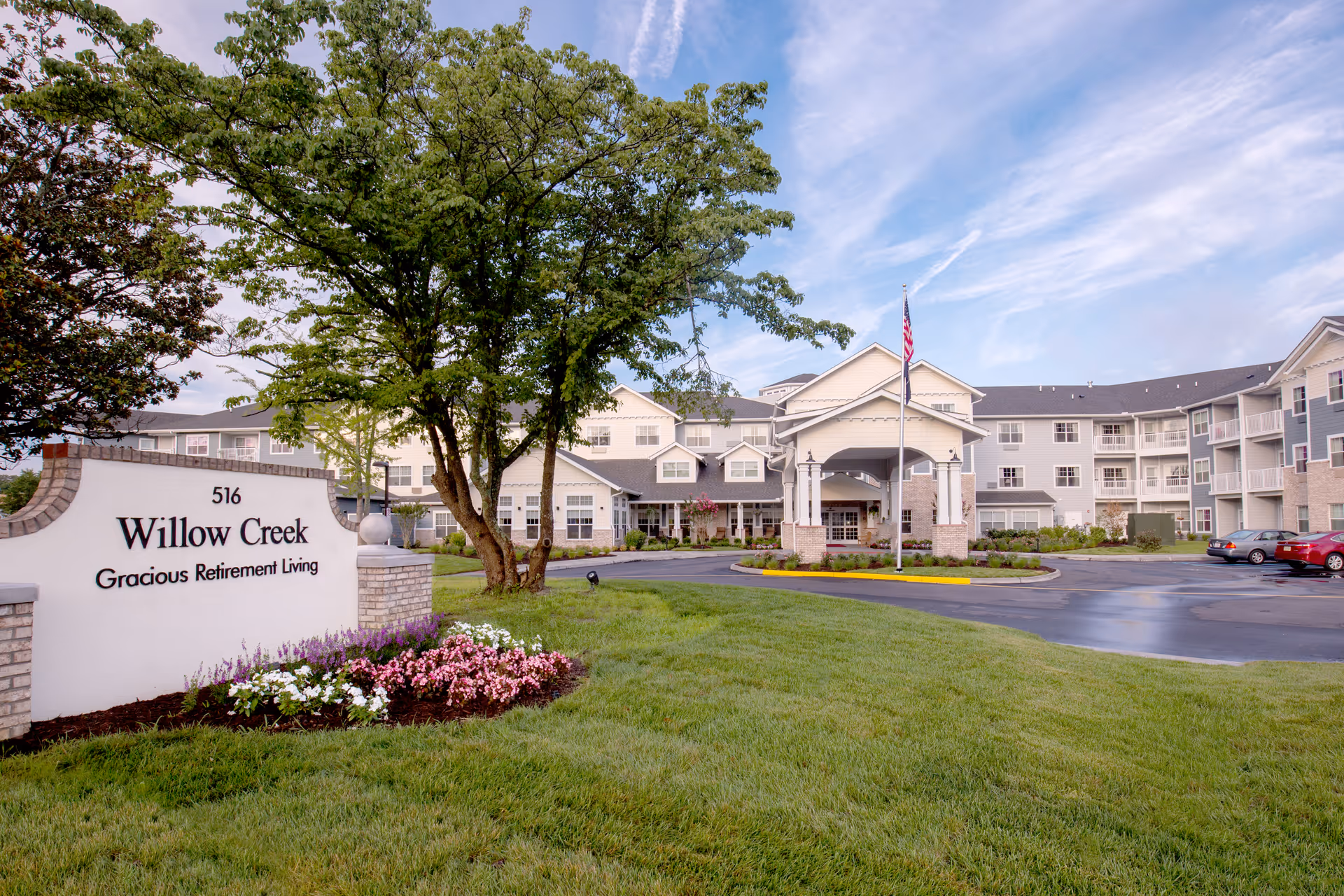 Exterior view of Willow Creek Gracious Retirement Living facility showing a large building with multiple windows and balconies, a covered entrance with an American flag in front, a well-maintained lawn, trees, and a flower bed near the entrance sign.