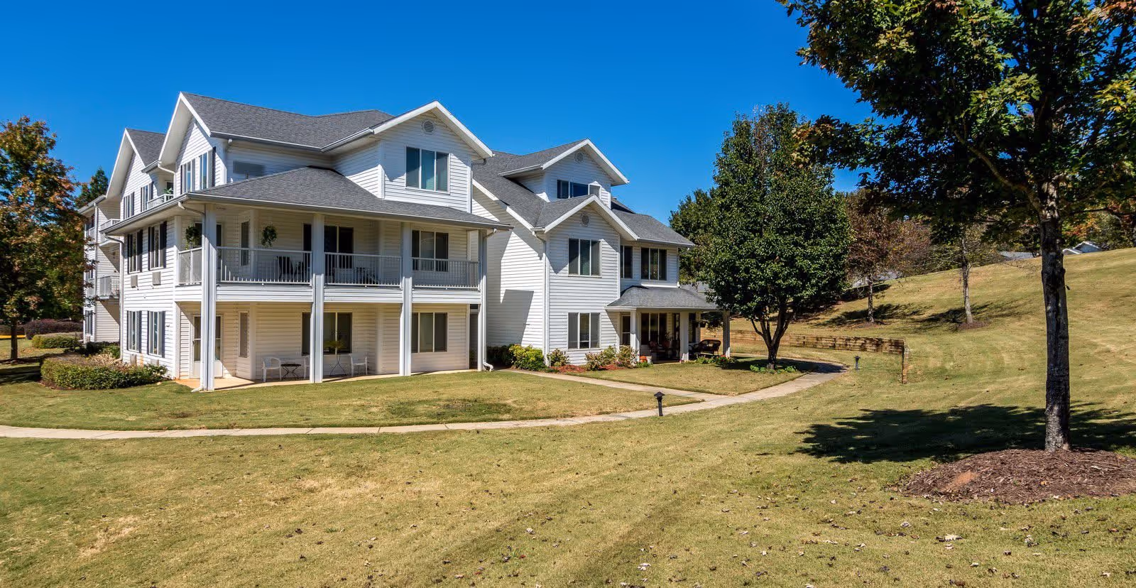 Exterior view of a multi-story white residential building with balconies, surrounded by a well-maintained lawn, trees, and a clear blue sky.