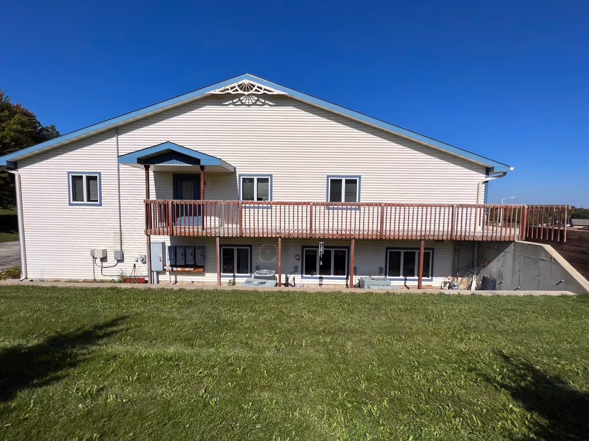 Front view of a two-story light-colored building with a wooden balcony and a grassy lawn under a clear blue sky.