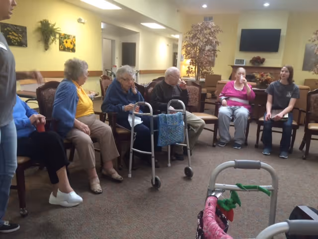 A group of elderly people sitting in a circle in a common area of a senior living facility, some using walkers. A younger woman is sitting with them, engaging in conversation. The room has warm lighting, chairs arranged in a circle, and a television mounted on the wall in the background.