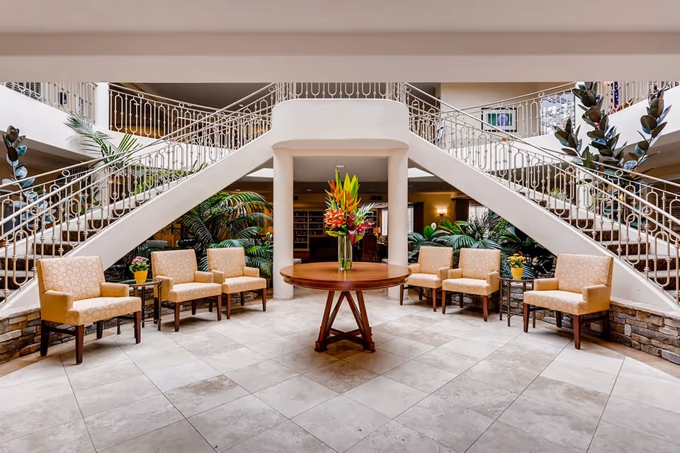 A spacious and elegant interior common area with a central round wooden table holding a vase of colorful flowers. The area is flanked by two symmetrical staircases with ornate white railings leading to an upper floor. Beige upholstered chairs are arranged in a semi-circle on both sides of the table, with small side tables holding potted plants. The floor is tiled with large light-colored tiles, and there are green plants placed around the space, creating a welcoming atmosphere.
