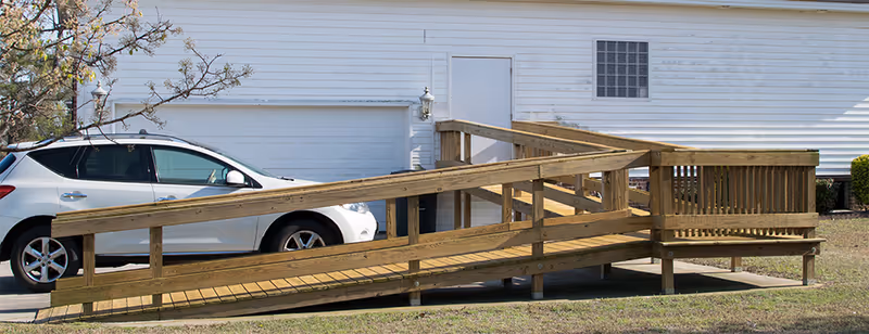 A wooden wheelchair ramp leading to a white building with a closed garage door and a white door. A white car is parked next to the ramp, and there is a small tree with light pink blossoms on the left side of the image.