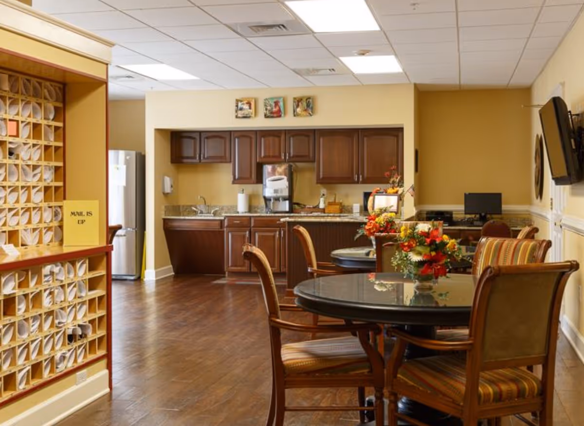 Communal dining area with round tables and chairs, floral centerpieces, and a kitchenette with wooden cabinets in the background.