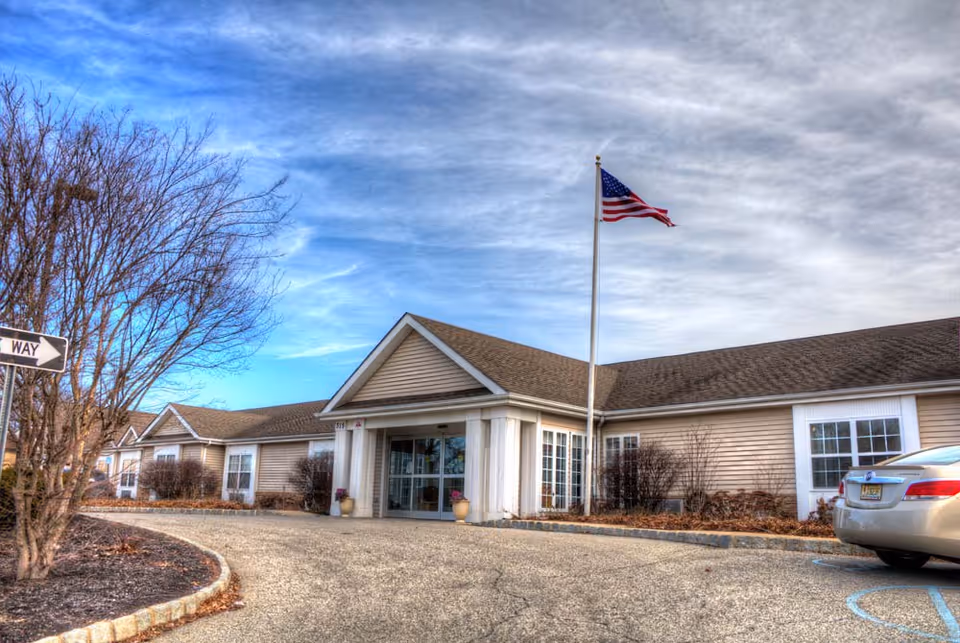 Exterior view of Merion Gardens Assisted Living facility showing a single-story building with beige siding, a peaked roof, and a covered entrance. An American flag is flying on a flagpole near the entrance. There is a car parked in a handicapped parking space to the right, and leafless trees and shrubs surround the building under a partly cloudy sky.