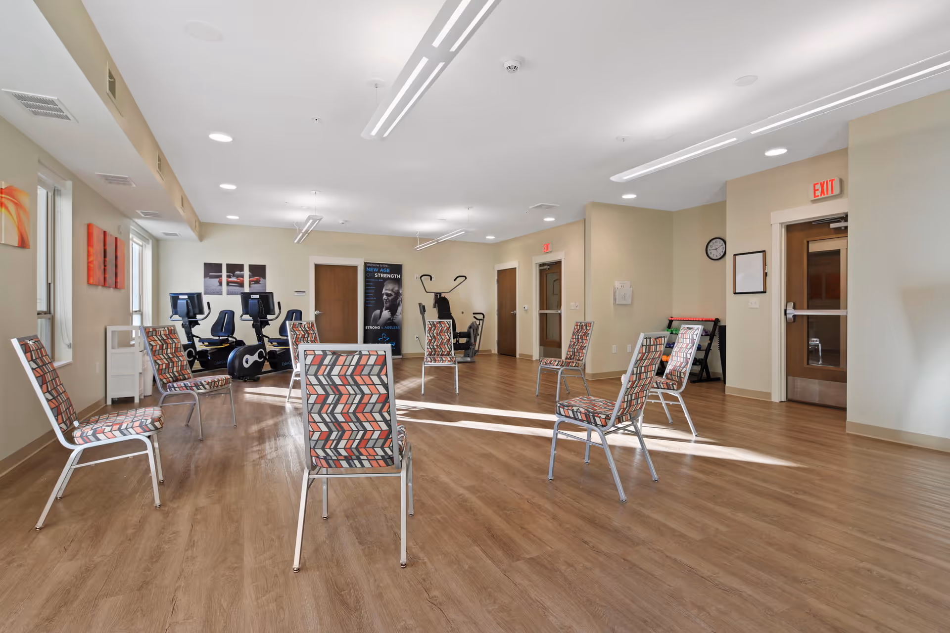 A spacious exercise room with wooden flooring and several patterned chairs arranged in a circle. There are exercise bikes and other fitness equipment against the far wall. The room has beige walls, multiple doors, and ceiling lights. A poster on the wall reads 'New Age Strength'.