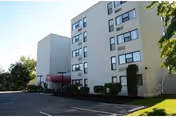 Multi-story senior living building with a red-canopied entrance, parking lot, and landscaping.