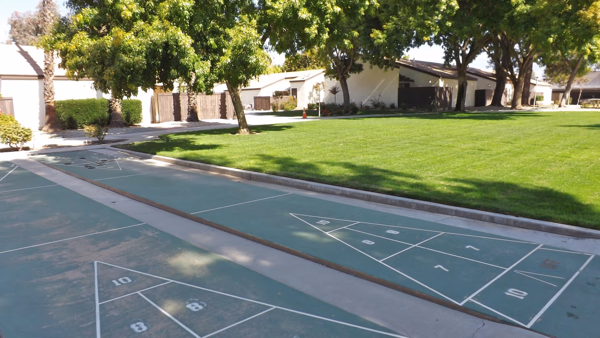 Outdoor shuffleboard courts beside a green lawn with trees and single-story buildings in the background.
