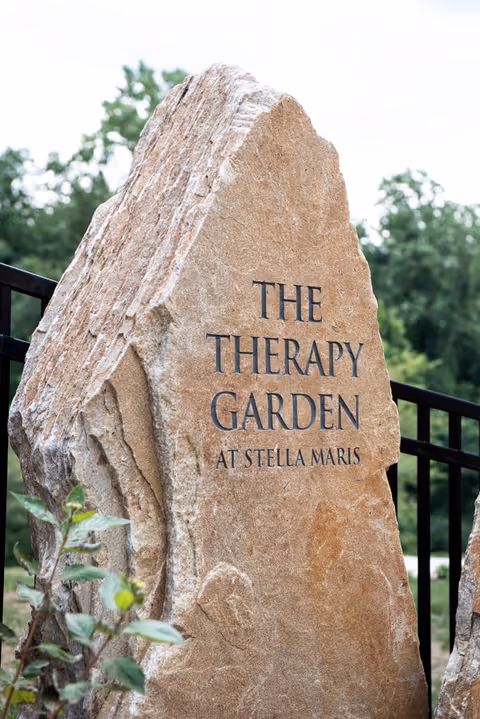 A large natural stone with engraved text that reads 'THE THERAPY GARDEN AT STELLA MARIS' set outdoors with greenery and a black metal fence in the background.