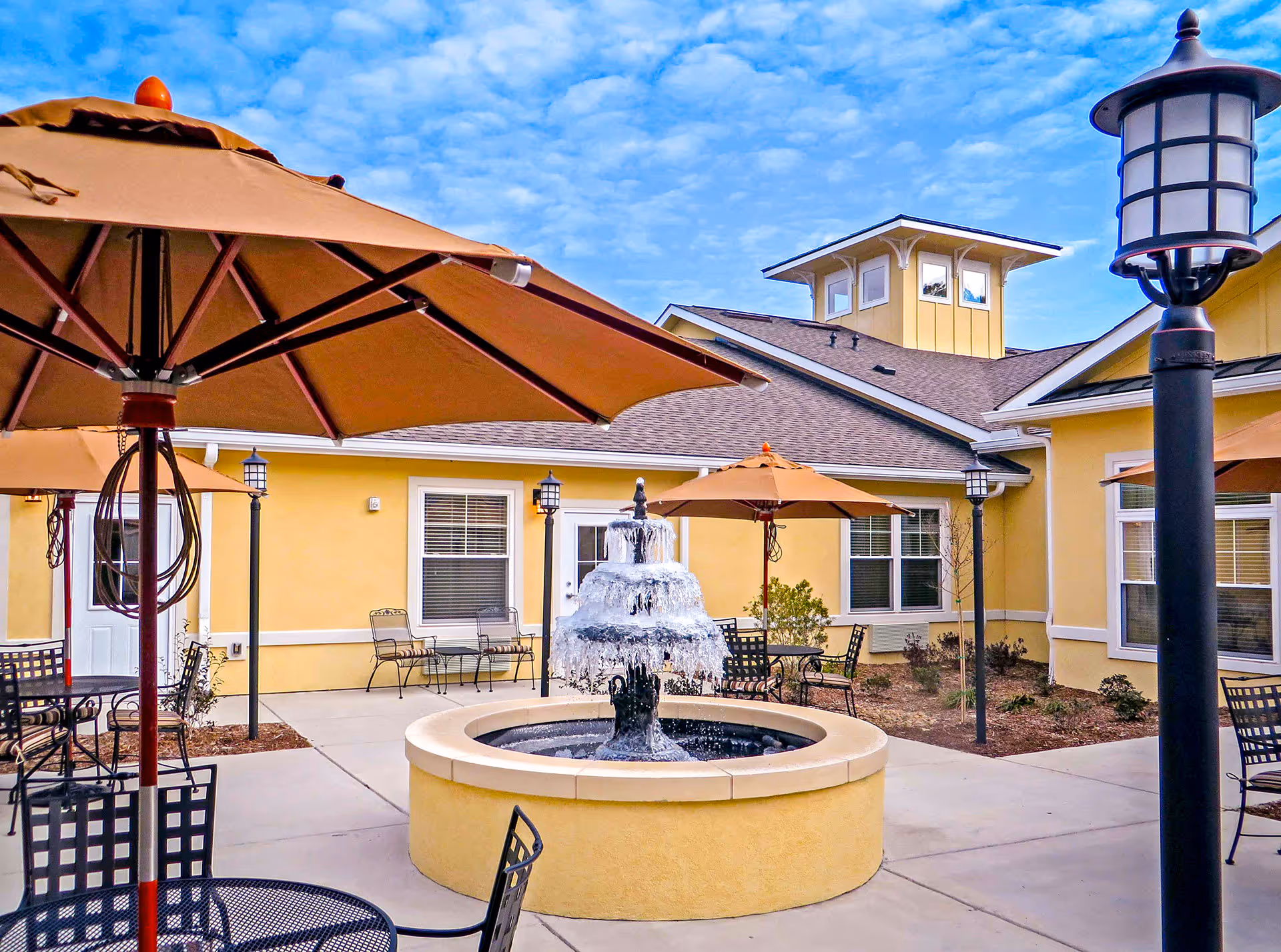 Outdoor courtyard area at The Addison of Bluffton featuring a central water fountain surrounded by patio tables with umbrellas and chairs. The building has yellow walls and multiple windows, with a clear blue sky overhead.